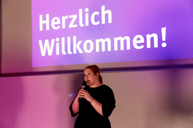 Kanzlerin Svenja Stepper spricht mit einem Mikrofon vor einer Leinwand, auf der groß „Herzlich Willkommen!“ steht. Die Szene ist in violettes Licht getaucht.