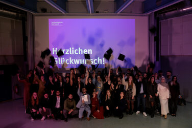 Die Absolvent*innen werfen ihre Absolventenhüte in die Luft, während im Hintergrund „Herzlichen Glückwunsch!“ auf der Leinwand steht. Die Szene feiert den Abschluss in festlicher Stimmung.