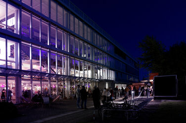 Das Gebäude des Fachbereichs ist abends in violettem Licht erleuchtet, und Menschen feiern drinnen und draußen. Vor dem Gebäude stehen Gruppen im Gespräch und einige Liegestühle.