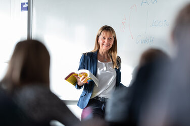A professor stands smiling at the front of the room in front of a white board with notes. She holds a book in her hand and looks at her students sitting in the room. Only a few of the students are vaguely recognizable from behind. __ <br>A female professor stands in front of the room smiling in front of a white board with notes. She holds a book in her hand and looks at her students who are sitting in the room. Only a few of the students are out of focus from behind.