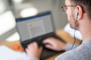Photo of a man at laptop in home office with headphones in ear. __ Man at laptop in home office with headphones in ear.