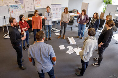 Photo of employees standing in a circle surrounded by flipcharts in a seminar. __ In a seminar, employees stand in a circle surrounded by flipcharts.