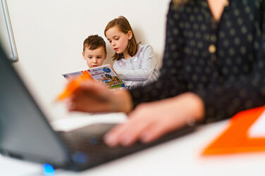 Photo of two younger children reading together. In the foreground you can see a laptop on the desk out of focus and someone who is working. __In the foreground you can see a laptop on the desk out of focus and someone who is working. The focus is on the children reading in the background.