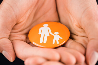 Photo of two hands lying on top of each other and holding a button with an icon in the camera. The icon represents a man and a woman in one part of the icons - as a split person, so to speak - a child is being held by one hand. __ <br>Two hands are on top of each other and hold a button with an icon in the camera. The icon represents a man and a woman in one part of the icons - as a kind of divided person - a child is held on one hand.