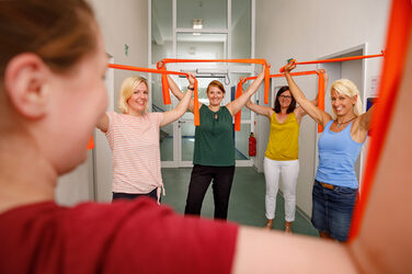 Photo of employees taking part in the break express in the corridor with orange Therabands. They hold the bands over their heads with their arms to stretch. __ Female employees take breaks in the hallway with orange Thera bands that they hold over their heads with their arms to stretch.