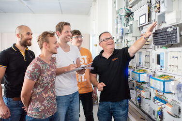 Photo of a professor standing in front of an electrical engineering test wall and pointing at a device. Next to him are four smiling students, one of whom is taking notes. __ A professor stands in front of an electrical engineering test wall and points to a device. Next to him are four smiling students, one of whom is taking notes.