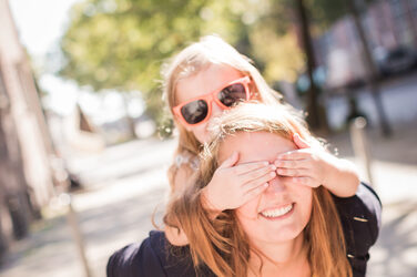 Photo of a young woman piggybacking a little girl with sunglasses. The girl holds the woman's eyes.__Photo of a young woman piggybacking a little girl with sunglasses. The girl holds the woman's eyes.
