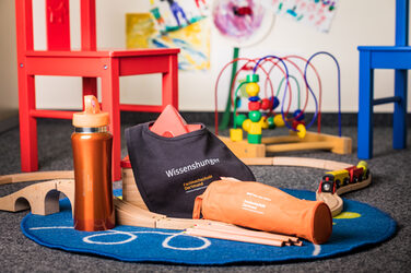 Photo of a still life on the floor consisting of; water bottle, wooden train set, bib labeled "hunger for knowledge" and the word mark of Fachhochschule Dortmund, pencil case and in the background chairs and another toy.__Photo of a still life on the floor consisting of; water bottle, wooden train set, bib labeled "hunger for knowledge" and the word mark of Fachhochschule Dortmund, pencil case and in the background chairs and another toy.