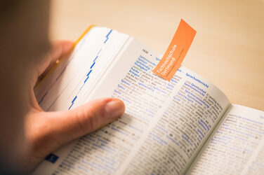 Photo of an open dictionary held in someone's hand. The open page is marked with an orange slip of paper with the word mark of Fachhochschule Dortmund __Photo of an open dictionary held in someone's hand. The open page is marked with an orange slip of paper with the word mark of Fachhochschule Dortmund.