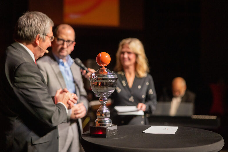 Foto von einem Stehtisch auf dem ein Pokal steht. Im Hintergrund verschwommen stehen eine weibliche und zwei männliche Personen. Die weibliche Person hält ein Mikrofon in der Hand.