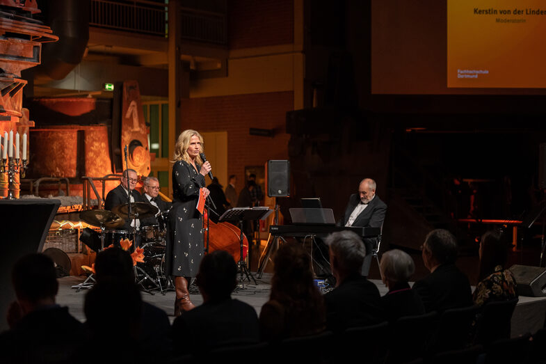 Foto einer weiblichen Person. Diese steht auf einer Bühne und spricht in ein Mikrofon zum Publikum, welches vor der Bühne sitzt. Im Hintergrund der Bühne sitzen drei männliche Personen einer Band vor ihren Musikinstrumenten.