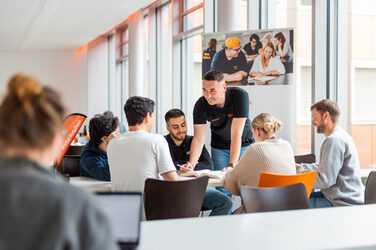 Auf dem Bild sieht man 5 Personen an einem Tisch sitzen, eine Person steht und zeigt auf etwas auf dem Tisch. Die restlichen Personen hören Ihm zu. Im Vordergrund erkennt man unscharf eine Person, die an einem Laptop arbeitet.