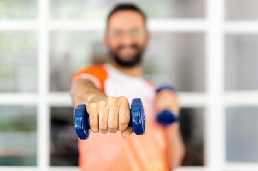 Es ist im Vordergrund eine Hand mit einer Hantel zu sehen, die in die Kamera gestreckt wird. Im Hintergrund sieht man unscharf die Person, die die Hantel hält in einem FH Dortmund Laufshirt