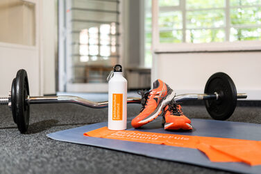 Verschiedenes Sportequipment auf einer Sportmatte. Eine Langhantel, orangene Sportschuhe. Dazu eine Wasserflasche und mit dem Fh Dortmund Logo sowie das FH Dortmund Theraband