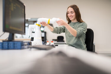Photo of a student sitting at a computer desk and holding the arm of a Schrenkarm robot with her hand.
