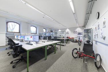 A picture of the Lightweight Construction Technology Center with work tables on which monitors are placed. In the center of the lab and on the right wall are display cases with exhibits from vehicle development. At the front right is a recumbent tricycle, at the back of the lab are an electric go-kart and a Pedersen bicycle with electric drive.