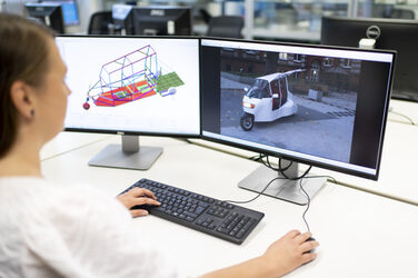 A student is sitting in front of two monitors. The right-hand monitor shows a 2-seater electric city vehicle with gullwing doors in vehicle class L2e with bodywork. A 3D FEM model of the vehicle can be seen on the left-hand monitor.