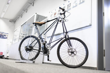 A Pedersen bicycle made of CFRP and aluminum with an electric drive via a mid-mounted motor stands in front of a wall in the Lightweight Technology Center. There is a shelf in the background and a whiteboard on the wall.