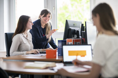 Photo of 3 women at desks. On the left of the picture is a young woman sitting at a desk, to her left is a woman standing next to her pointing at the monitor. On the right, out of focus, is a woman with her back to the camera, writing something
