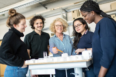 Photo of a professor, surrounded by her students, stands behind an architectural model and points with a pen at a part to explain something to her students. __ A professor, surrounded by her students, stands behind an architectural model and points with a pen at a part to explain something to her students.