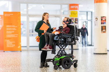 Photo of an employee in a wheelchair raised to the eye level of the colleague standing next to him. Both colleagues are talking in the foyer. __ <br>Employee sits in a wheelchair, which has been raised at eye level with the colleague standing next to him. Both colleagues are talking in the foyer.