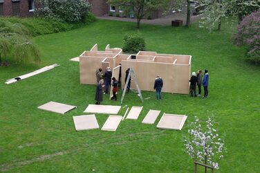 Der Grundriss ist festgelegt und einige Wandteile sind angebracht, so dass Strukturen eines Labyrinths sichtbar sind. Die Studierenden des Workshops stehen verteilt rundherum, umgeben von weiteren Bauteilen, Dachlatten und einer Leiter.