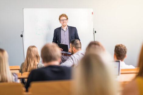 Photo of a lecturer in a lecture hall standing in front of a whiteboard, laughing. In the foreground, several seated students are blurred and seen from behind.