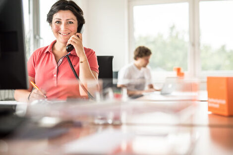 Photo of an employee sitting at her desk talking on the phone and smiling at the camera. In the background, a man is working on his laptop.