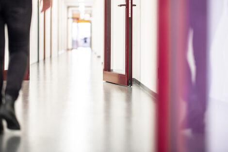 Photo of a long corridor with a red door in the foreground. A person's legs are on the left edge of the picture.