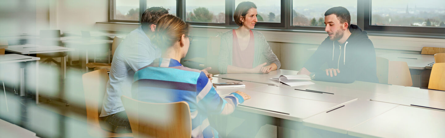Photo through a window pane into a seminar room where four students are sitting at a table and exchanging ideas.