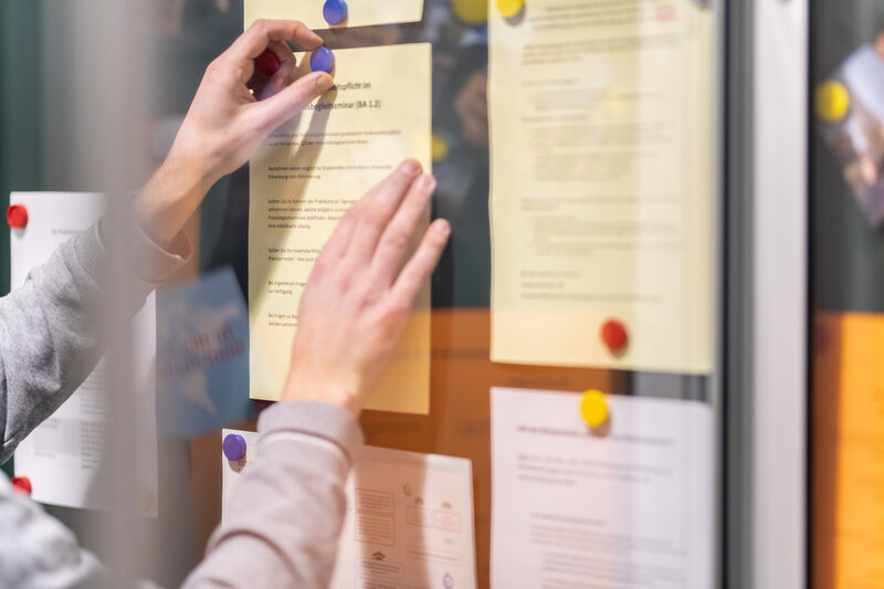 Photo of a display case with several notes pinned inside. One person pins up another piece of paper with magnets.