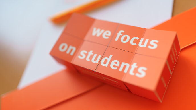 Photo of an opened folding cube with the imprint "we focus on students" lies on documents. __ Opened folding cube with the imprint "we focus on students" lies on documents.