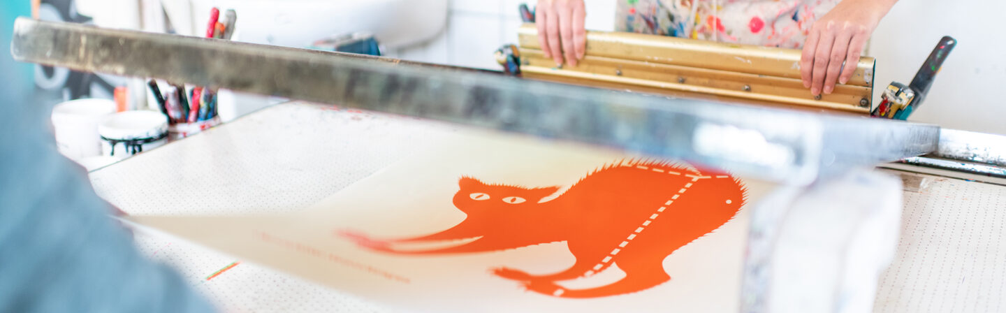 Photo of two people working together to create a screen print of an orange cat. Only the upper body and arms of the people can be seen.