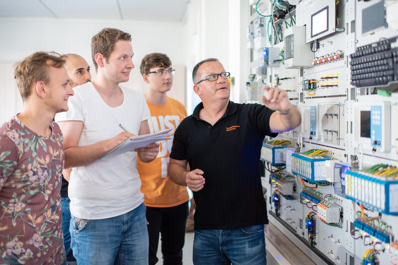 Photo of a professor with his students. They are standing in front of an electrical engineering test wall, the professor is pointing at a device. One of the students is taking notes.