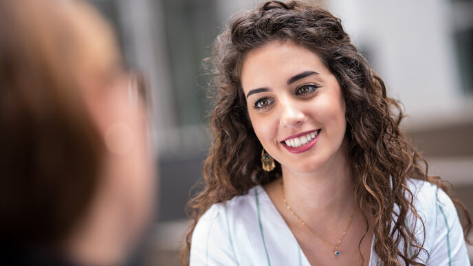 Closeup of a young woman with dark curly hair smiling at a woman sitting across from her.__Closeup of a young woman with dark curly hair smiling at a woman sitting across from her.