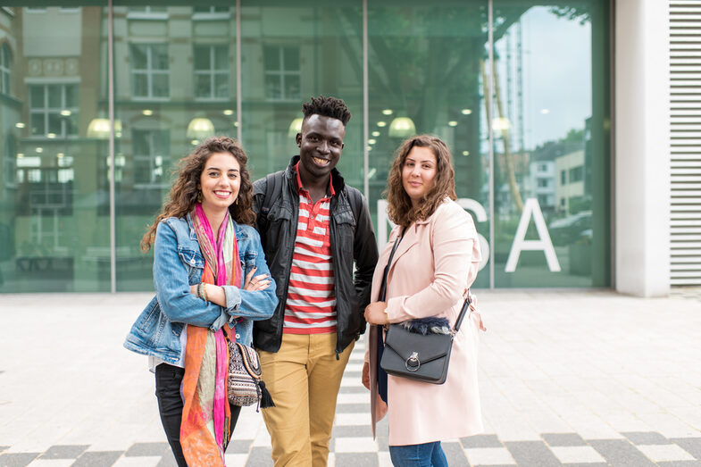 Photo of two female and one male students standing side by side in front of the canteen building. They laugh at the camera.__Two female and one male students stand side by side in front of the canteen building. They laugh at the camera.