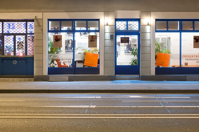 Photo of the university's project office on site from the outside: A house wall with large blue-framed windows through which you can see into the building