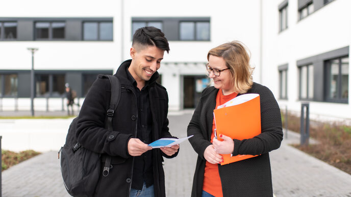 Photo of an employee and a student standing in front of a campus building. She is holding an orange folder under her arm, he has a flyer in his hand. They both look at the flyer and laugh.