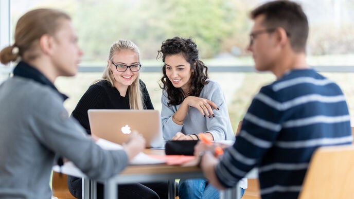Photo of four students at a group table. The focus is on two female students sitting next to each other and looking at a laptop. Two other students are engrossed in a conversation.