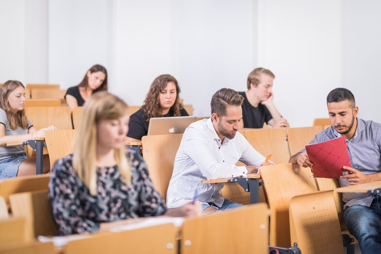 Photo of rows of seats in the lecture hall with several students. Everyone is concentrating on their desks and writing, reading or typing on their laptops.