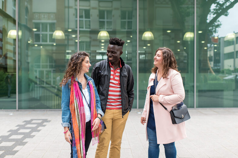 Photo of two female and one male students standing side by side in front of the canteen building, they laugh at each other. They laugh at each other.__Two female and one male students stand side by side in front of the canteen building, they laugh at each other.