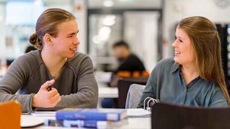 Photo of a female student and a male student sitting next to each other at a table, they laugh at each other. Books are on the table.__Student sit next to each other at a table, they laugh at each other. Books are on the table.<br><br>