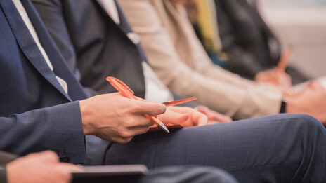 Photo of a row of people sitting at an event. A man holding pen and material in hand. __ Row of people sitting at an event, man holding pen and material in hand.