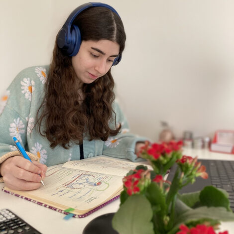 Mrs. Saleh writing in a notebook in front of her notebook sitting at a table. She wears blue headphones on her head.<br>In the foreground, blurred to see, a flower.
