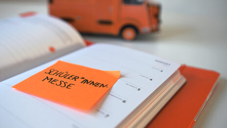 Photo of an orange Post-It with the word "Schüler*innen-Messe" stuck to a calendar. In the background is a pencil box on the table.