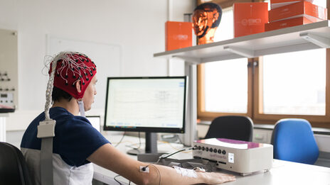 Photo of a test person with EEG cap and neuroreceptor on elbow in front of a measurement screen.