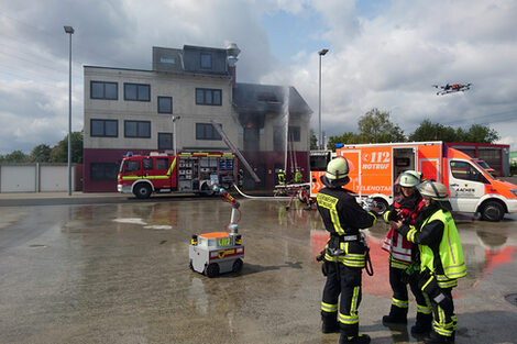 Die Feuerwehr löscht einen brennenden Dachstuhl, im Vordergrund steht ein Roboter. Feuerwehrleute steuern zudem eine Drohne.