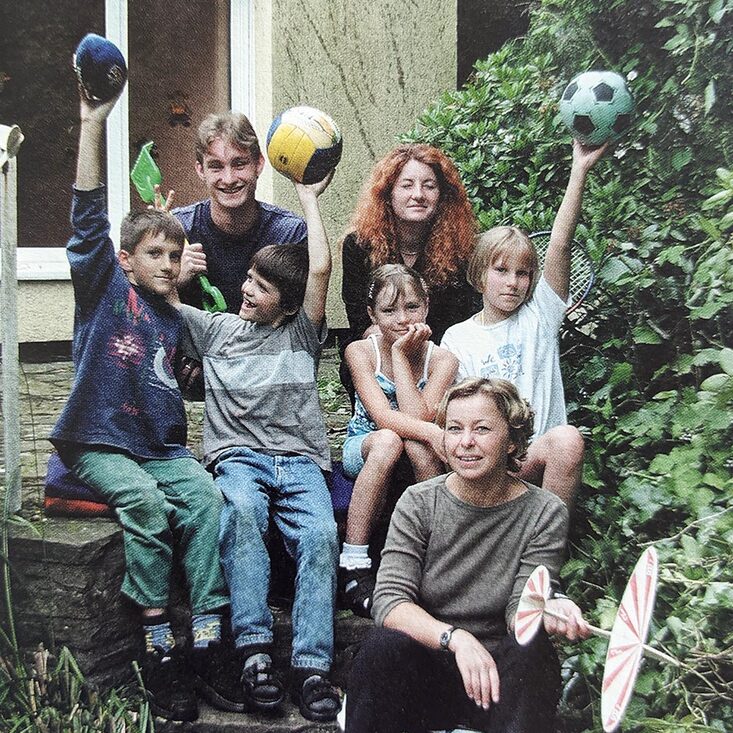 Vier Kinder und drei Erwachsene sitzen auf einer Außentreppe vor einem Gebäude. Die Kinder strecken Bälle in die Höhe.