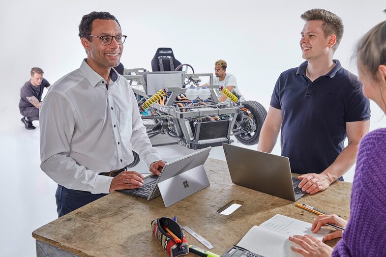 Im Vordergrund stehen drei Menschen an einem Tisch: Ein älterer Mann links an seinem Laptop, der lächelt. Rechts von ihm ein junger Mann, ebenfalls am Laptop, der zu dem anderen Mann blickt. Von schräg hinten sieht man zudem eine junge Frau, die auf einen Notizblock vor sich blickt. Im Hintergrund arbeiten zwei junge Männer an einer Fahrzeugkarosserie.