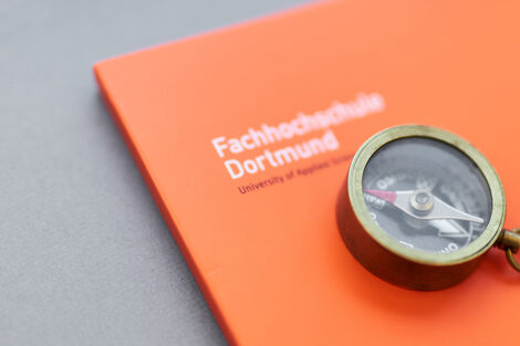 Photo of a small compass with a chain on it, lying on an orange folder of the Fachhochschule Dortmund.